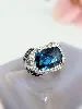 bague cocktail centrée d'une topaze facetée serti clos entourée de petits diamants et de saphirs sur le panier or 750 millième (18
